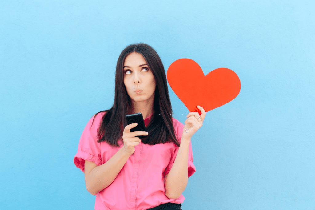 A young lady holding smartphone and a heart in her hands and thinking about dating apps
